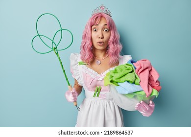 Impressed Shocked Housemaid Collects Colorful Laundry In Plastic Basin Holds Carpetbeater Wears White Dress Does Daily Cleaning Isolated Over Blue Background. Housework And Housekeeping Concept