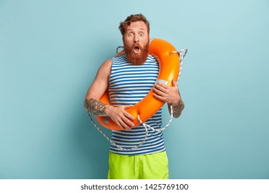 Impressed shocked red haired lifeguard works at beach, uses lifebuoy for saving people, wears striped sailor vest and green shorts, stands indoor, feels embarrassed. Omg and water activities concept