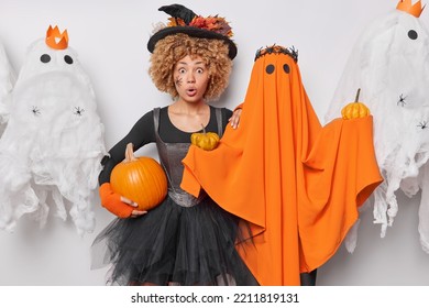 Impressed Shocked Woman With Curly Hair Wears Black Dress And Hat Holds Pumpkin Poses Near Orange Ghost Stares Amazed At Camera Believes In Magic Celebrates Hallooween Comes On Costume Party