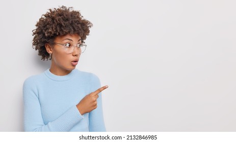 Impressed Stupefied Young Woman Points Index Finger Away Shows Place For Your Advertisement Reacts On Big Discounts Wears Eyeglasses And Casual Jumper Isolated Over White Background. Omg Look There