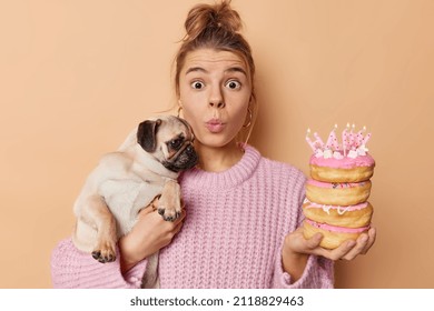 Impressed Suprised Young Woman Stares With Widely Opened Eyes Holds Pug Dog On Hands Poses With Festive Tasty Doughnuts Keeps Lips Folded Wears Knitted Sweater Isolated Over Brown Background