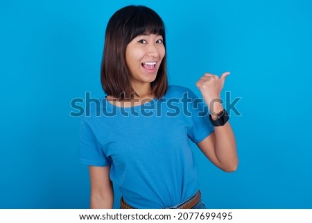Impressed young asian woman wearing blue t-shirt against blue background point back empty space