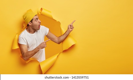 Impressed young man points away, shows direction somewhere, gasps from wonderment, has anxious worried facial expression, dressed in white t shirt and headgear, poses in torn hole of paper wall