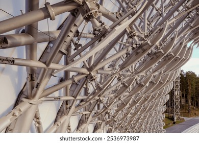 An impressive array of metallic beams and supports showcases engineering brilliance, illuminated by the sun, surrounded by serene greenery in a wooded setting.