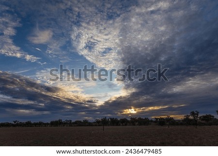 An impressive sunset sky with swirls of clouds and a moody and awe inspiring ambience with grass and trees in the foreground in the town of Cunnamulla in Queensland, Australia.