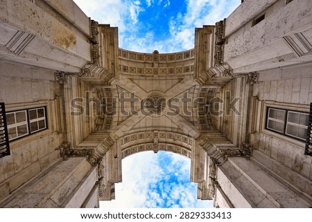 An impressive symmetrical looking-up perspective capturing the detailed vaulted ceiling and massive limestone columns of the Rua Augusta Arch (Arco da Rua Augusta) in Lisbon, Portugal. The architectural shot showcases the intricate stone medallions, geometric relief carvings, and classic windows on the interior walls of this historic triumphal arch. Framed perfectly by the monumental structure, a vibrant blue sky with soft white clouds peaks through the top and bottom openings