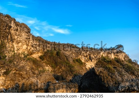 Impressive wide angle view of a massive, textured rocky cliff face along a tropical coastline. Giant boulders line the shore next to clear ocean water under a beautiful bright blue sky.