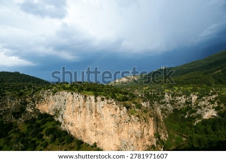 An impressive wide panoramic view of a massive sheer rocky cliff face rising above a lush green forest under a dark stormy sky.