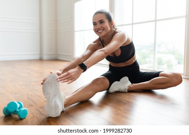 Improve Flexibility. Positive cheerful young female stretching leg and hamstring muscles sitting on the floor after or before workout, wearing sportswear, wristwatch and white sneakers, free space
