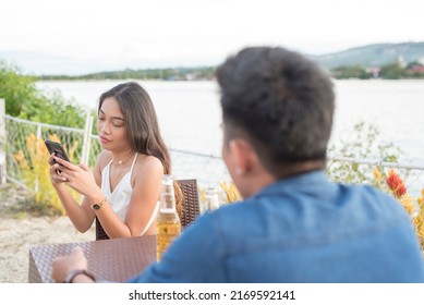 An Inattentive Young Woman Checks Her Phone While Ignoring A Man Talking To Her At The Moment. Being Rude, Bored Or Simply Uninterested In Her Date. A Failed First Date.