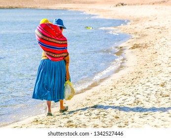 Incan Woman With Baby In The Colorful Scarf On A Back. Walks On The Beach Of Island Of The Sun At Titicaca Lake, Bolivia, South America.