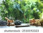 Incense stick smoldering in holder, burning candles, dry flowers and Tibetan singing bowl on wooden table outdoors