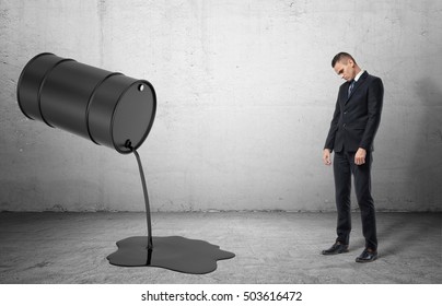 An Inclined Barrel With Black Liquid Pouring Out Of It On A Grey Floor And A Dissapointed Sad Businessman Full Height, On The Grey Background. Business And Finance. Oil And Gas Industry. Failures And