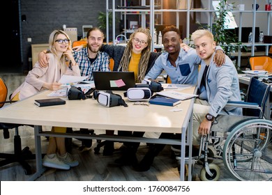 Inclusion in work, business team concept. Handsome male coworker on wheelchair with his creative business colleagues in meeting room at creative office, smiling, looking at camera, hugging each other