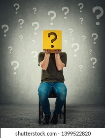Incognito young man seated on a chair holding a yellow box with question mark instead of head. Introvert person anonymity concept hiding identity behind a mask. Social issue, shy guy covering face.