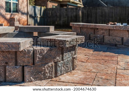 Incomplete paver patio hardscape with a stone fire pit and seating wall under construction in a fenced suburban backyard