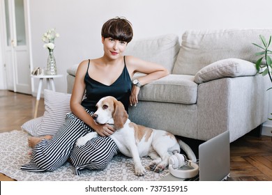 Incredible Brunette Lady Wears Elegant Wristwatch Posing Beside Sofa Stroking Cute Beagle. Indoor Portrait Of Blissful Dark-haired Girl Sitting On Carpet With Dog And Looking To Camera With Smile.