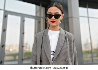 Incredible stylish woman with dark hair and red lips wearing golden earrings and grey jacket posing at camera outdoor against glass building. 