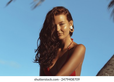 Incredibly beautiful woman in a red bikini smiles warmly under a bright blue sky, her wavy hair cascading over her shoulder. The tropical palm leaves in the background add to the serene and relaxed - Powered by Shutterstock