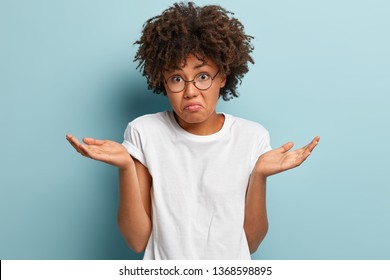 Indecisive Dark Skinned Girl Spreads Palms With Confused Expression, Wears Casual Mock Up T Shirt, Frowns Face In Uncertainty, Has Afro Haircut, Isolated Over Blue Background. Woman Faces Trouble