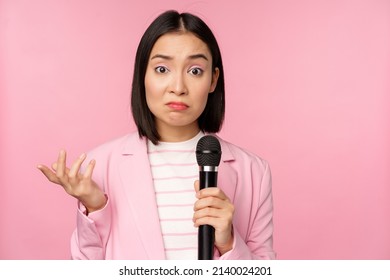 Indecisive, Nervous Asian Business Woman Holding Mic, Shrugging And Looking Clueless, Standing With Microphone Against Pink Background, Wearing Suit