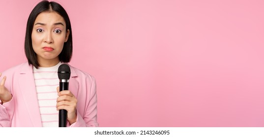 Indecisive, Nervous Asian Business Woman Holding Mic, Shrugging And Looking Clueless, Standing With Microphone Against Pink Background, Wearing Suit