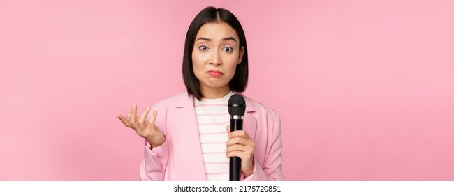 Indecisive, Nervous Asian Business Woman Holding Mic, Shrugging And Looking Clueless, Standing With Microphone Against Pink Background, Wearing Suit