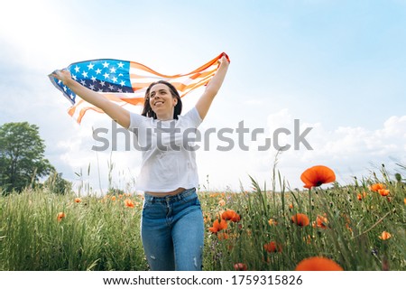 Independence day. Happy cheerful young caucasian girl is jogging across poppy field holding usa flag 