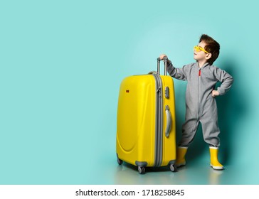 Independent kid boy in gray overall, yellow rubber boots and sunglasses stands at big roller bag wheeler looks forward up at copy space over blue background. Travelling concept