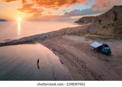  Independent road trip concept: Campervan on Abandoned Beach against beutiful Sunset,  Woman bathing in the Sparkling sea. Outdoor nomad lifestyle, van life holiday. Aerial photo. 