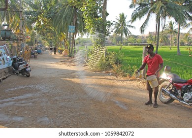 India, Hampi, 02 February 2018. The Main Street Of The Village Of Virupapur Gaddi. Guesthouses Overlooking The Rice Fields. An Indian Man Sprinkles Water On The Road. Drops Of Water In Flight.