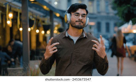 Indian Arabian male man teen gen z happy guy student businessman tourist in modern wireless headphones having fun listen relax music song moving dance gesticulate dance outdoors night city town street - Powered by Shutterstock