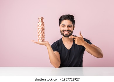Indian asian bearded young man holding or presenting copper water bottle against pink background while sitting at table