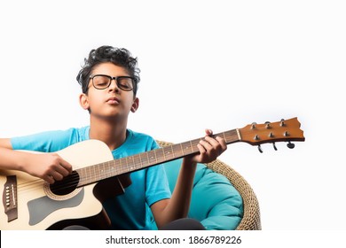 Indian asian boy playing acaustic guitar while sitting against white background or brick wala on chair