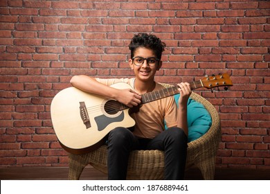 Indian asian boy playing acaustic guitar while sitting against white background or brick wala on chair