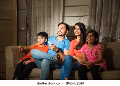 Indian asian family of four watching television or TV while sitting on sofa or couch