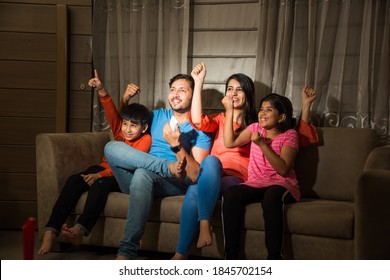 Indian Asian Family Of Four Watching Television Or TV While Sitting On Sofa Or Couch