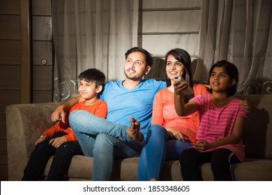 Indian Asian Family Of Four Watching Television Or TV While Sitting On Sofa Or Couch