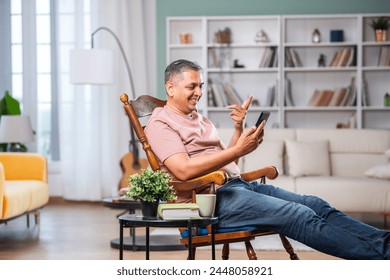 Indian asian man in 40s with grey hair using smartphone at home, sitting on rocking relaxing chair - Powered by Shutterstock