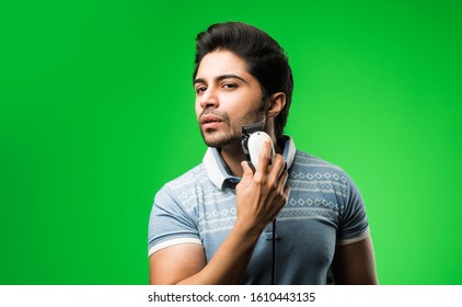 Indian / Asian Man Shaving Beard With Electric Trimmer Or Clipper Standing Isolated Over Green Background