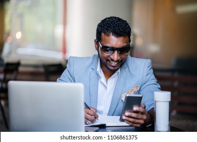 An Indian Asian man is sitting at a coffee shop and is glancing at his phone and writing in his notebook. His laptop is nearby and he is smiling as he does his work. He is dressed in a casual suit. 