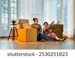 Indian Asian middle-aged married couple sitting on the floor of their new home surrounded by moving boxes, using a laptop to work on interior design plans, happily settling into their new apartment