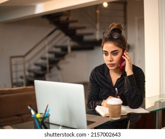 Indian Asian Modern young Businesswoman or corporate female Entrepreneur writing notes in a diary while talking or having a conversation on a Mobile Phone in an interior house setup. work from home co