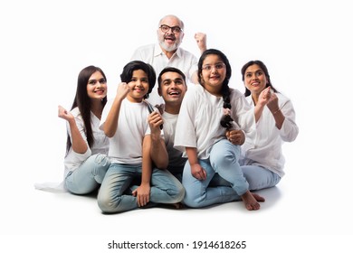 Indian Asian Multigenerational Family Of Six Watching TV Together At Home And Having Fun , Sitting On Floor Against White Background