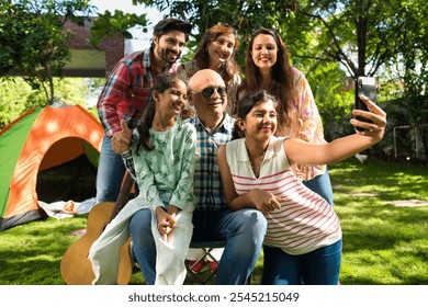 Indian Asian multigenerational family of six camping outdoors, enjoying music, singing, and capturing moments with a selfie, creating joyful memories together - Powered by Shutterstock