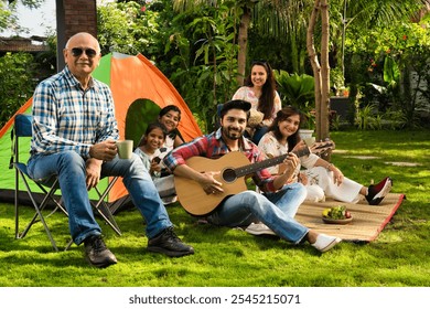 Indian Asian multigenerational family of six camping outdoors, enjoying music, singing, and capturing moments with a selfie, creating joyful memories together - Powered by Shutterstock