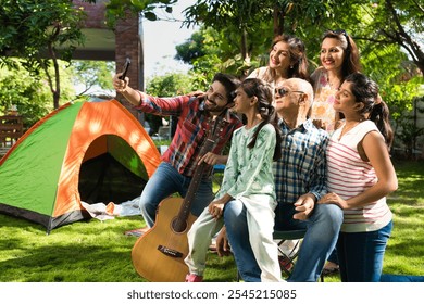 Indian Asian multigenerational family of six camping outdoors, enjoying music, singing, and capturing moments with a selfie, creating joyful memories together - Powered by Shutterstock