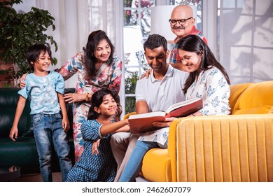 Indian asian multigenerational family watching photos in album or reading book while sitting on sofa together - Powered by Shutterstock