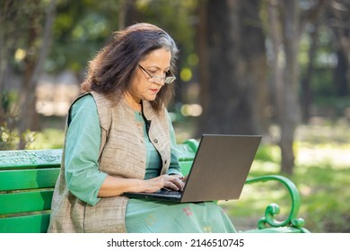 Indian asian senior woman sitting on bench using laptop at park. Old or mature people wearing eye glasses working on computer, modern technology concept.