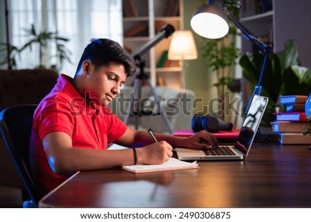 Indian Asian teenage boy studying using computer, headphones, smartphone for online class, homework
